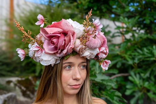 May include: A floral crown featuring large, dusty rose-colored roses, white flowers, and delicate pink accents. The crown is set against a backdrop of green foliage and a woman's face.