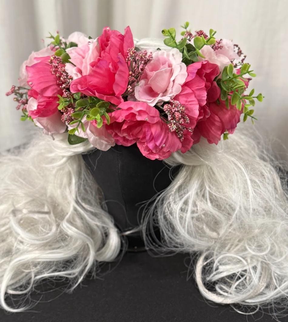 May include: A floral crown featuring vibrant pink and light pink flowers, accented with green foliage and small pink buds. The crown sits atop a black head form with long, flowing, white wig hair.