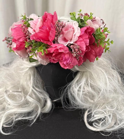 May include: A floral crown featuring vibrant pink and light pink flowers, accented with green foliage and small pink buds. The crown sits atop a black head form with long, flowing, white wig hair.